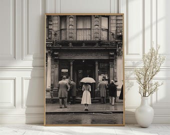 May include: Black and white framed print of a vintage street scene. A woman with an umbrella stands in front of a building with several people. The building has ornate architectural details and the word "FRIE" is visible.