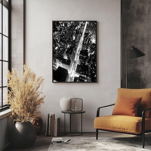May include: Black and white aerial photograph of a city at night. The photo shows a street with cars driving on it and buildings on either side of the street. The street is lit up by streetlights and car headlights.
