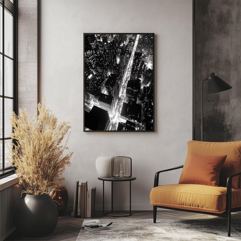 May include: Black and white aerial photograph of a city at night. The photo shows a street with cars driving on it and buildings on either side of the street. The street is lit up by streetlights and car headlights.
