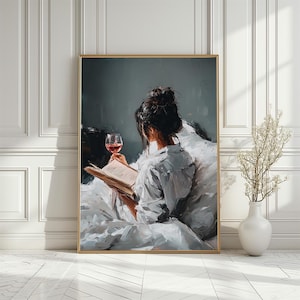 May include: A framed art print showing a person reading a book and holding a glass of red wine. The person wears a white shirt and sits on a bed with white bedding. The artwork has a soft, painterly style, with a muted colour palette.