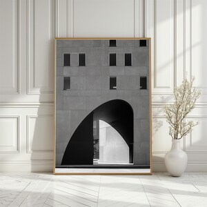 May include: Framed architectural print featuring a grayscale image of a modern building with an arched entrance. The building has multiple rectangular windows and a textured facade. The print is displayed in a gold frame, with a white vase and decorative branches in the foreground.