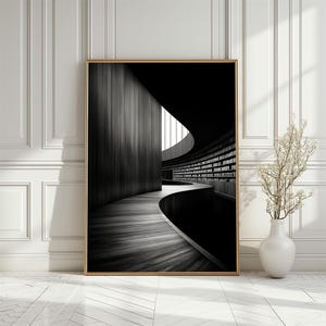 May include: A framed black and white photograph of a library interior. The image features a curved walkway leading into a library with bookshelves. The photograph is in a gold frame and is displayed against a white wall. A white vase with decorative branches is in the foreground.
