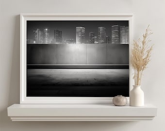 May include: A framed black and white landscape photograph featuring a city skyline at night above a concrete wall and paved ground. The print is displayed on a white shelf next to a small round ceramic vase and a tall white vase with dried grass.