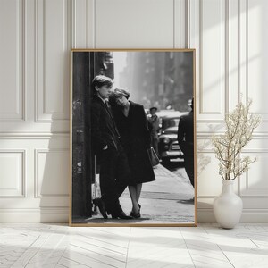 Könnte beinhalten: Schwarz-Weiß-Fotografie, gerahmt, eines Paares auf einer Stadtstraße. Der Mann trägt einen Anzug, die Frau einen Mantel. Ein Oldtimer und andere Figuren sind im Hintergrund zu sehen, was eine klassische Szene schafft.