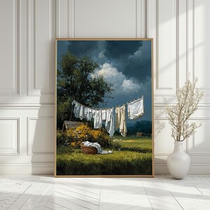 Op de afbeelding: Een ingelijst schilderij toont een landelijke scène met witte was die aan een waslijn hangt. Het kunstwerk toont een donkere, bewolkte lucht, een groen veld en een mand met kleding. Een boom en een klein gebouw zijn ook zichtbaar.