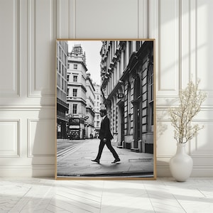 Könnte beinhalten: Ein Schwarzweißfoto eines Mannes, der eine Stadtstraße entlangläuft. Der Mann trägt einen Anzug und eine Krawatte. Die Straße ist von hohen Gebäuden gesäumt. Das Foto ist in einem goldenen Rahmen gerahmt.