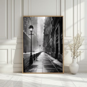 May include: A black and white photograph of a city street with tall buildings and a lamppost in the foreground. The street is empty and the buildings are lined up on either side of the street. The photograph is taken from a low angle, looking up at the buildings.