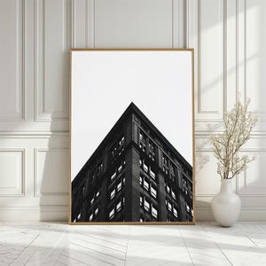 May include: A black and white framed wall art print featuring a low-angle perspective of a dark brick building corner against a bright white sky. The rectangular print is encased in a thin gold frame and displays a sharp architectural design.