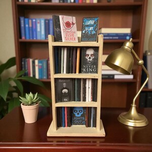 May include: A miniature wooden bookshelf filled with tiny books, including titles like "Flawless" and "The Never King." The bookshelf is on a dark wooden surface, with a small succulent in a pink pot and a brass desk lamp nearby.