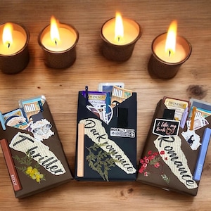 Op de afbeelding: Drie bruine dozen versierd met theezakjes, stickers en gedroogde bloemen. Elke doos heeft een ander genre van boeken erop geschreven: thriller, dark romance en romance. De dozen staan op een houten oppervlak met drie brandende kaarsen ervoor.