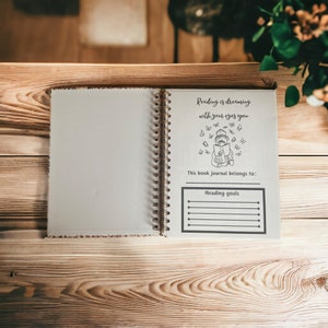 May include: An open journal with a white cover and a black and white illustration of a person reading a book with butterflies flying around them. The text on the page reads "Reading is dreaming with your eyes open." There is a section for "Reading goals" with lines to write on.