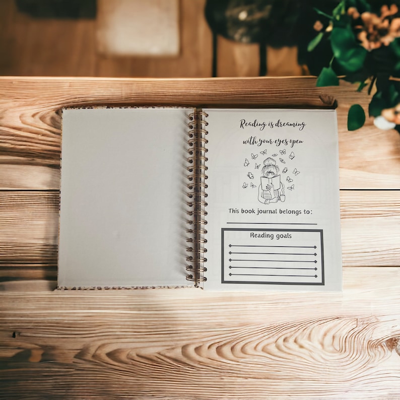 May include: An open journal with a white cover and a black and white illustration of a person reading a book with butterflies flying around them. The text on the page reads "Reading is dreaming with your eyes open." There is a section for "Reading goals" with lines to write on.