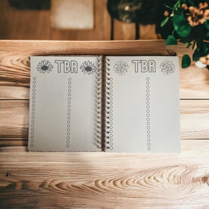 May include: A white spiral bound notebook with the text "TBR" written in black lettering. The notebook has a floral design on the cover and is open to a blank page with a list of circles.