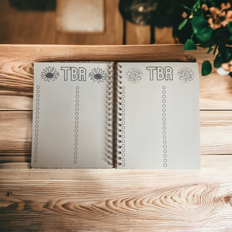 May include: A white spiral bound notebook with the text "TBR" written in black lettering. The notebook has a floral design on the cover and is open to a blank page with a list of circles.