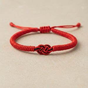 Op de afbeelding: Een rode verstelbare armband met een centraal knoopontwerp en zwarte details. De armband is gemaakt van een geweven rood koord en heeft een schuifknoopsluiting voor een aanpasbare pasvorm. Een eenvoudig maar elegant accessoire.