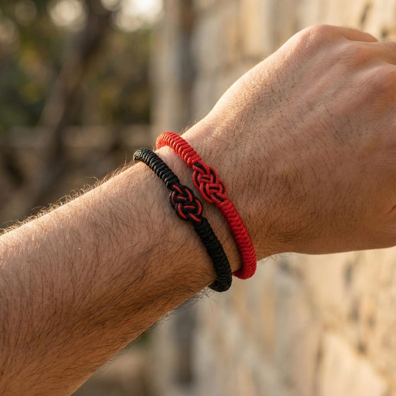 Op de afbeelding: Twee handgemaakte armbanden, &eacute;&eacute;n zwart en &eacute;&eacute;n rood. Beide armbanden hebben een geweven knoopontwerp in het midden. De armbanden worden om een pols gedragen, waarbij het ingewikkelde knoopwerk en de levendige kleuren worden benadrukt.