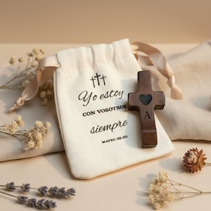 Könnte beinhalten: Ein dunkles Holzkreuz mit einem Herz-Ausschnitt und dem Buchstaben "A" liegt auf einem cremefarbenen Kordelzugbeutel. Der Beutel trägt den Text "Yo estoy con vosotros siempre" und einen Bibelvers. Das Kreuz wird mit Trockenblumen präsentiert.