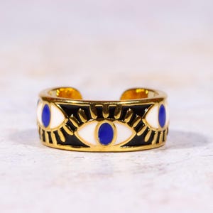 Puede incluir: Anillo dorado con una banda negra con dise&ntilde;os de ojos blancos y azules. El anillo tiene un dise&ntilde;o abierto y est&aacute; sobre un fondo neutro. Un accesorio &uacute;nico y elegante.