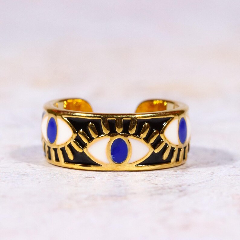 Puede incluir: Anillo dorado con una banda negra con dise&ntilde;os de ojos blancos y azules. El anillo tiene un dise&ntilde;o abierto y est&aacute; sobre un fondo neutro. Un accesorio &uacute;nico y elegante.