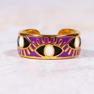 Puede incluir: Anillo ajustable dorado con una banda morada con un dise&ntilde;o de ojo. El anillo tiene detalles de ojos negros y blancos, con detalles dorados. El anillo es un accesorio de moda.
