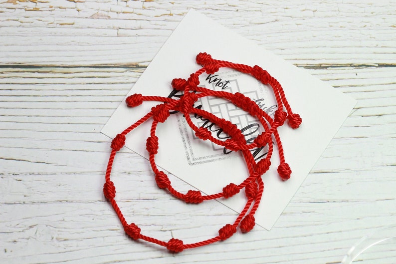 Op de afbeelding: Verschillende rode touwarmbanden met geknoopte details zijn gerangschikt op een witte kaart met tekst. De armbanden zijn gemaakt van een dun, flexibel materiaal en hebben meerdere knopen. De achtergrond is een lichtgekleurd, getextureerd houten oppervlak.