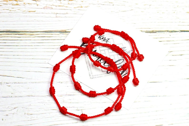 Op de afbeelding: Verschillende rode touwarmbanden met geknoopte details worden getoond op een wit houten oppervlak. De armbanden hebben meerdere knopen langs het touw en zijn gerangschikt in de buurt van een kaart met het woord "Knot" zichtbaar.