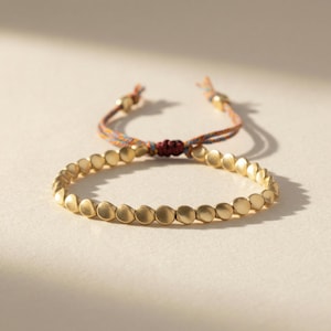 Könnte beinhalten: Ein goldfarbenes Armband mit einer Reihe kleiner, flacher, kreisförmiger Perlen. Das Armband ist mit einer mehrfarbigen Kordel mit Schiebeknotenverschluss verbunden. Die Kordel hat orange, blaue und rote Fäden.