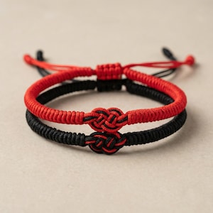May include: Two matching bracelets, one red and one black, are displayed. Each bracelet features a woven design with a knot in the center. The red bracelet is on top of the black bracelet. The background is a neutral color.