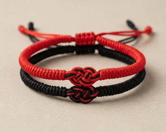 Puede incluir: Se muestran dos pulseras a juego, una roja y otra negra. Cada pulsera presenta un diseño tejido con un nudo en el centro. La pulsera roja está encima de la pulsera negra. El fondo es de color neutro.