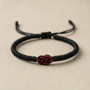 Op de afbeelding: Een zwart gevlochten armband met een rood knoopontwerp in het midden. De armband heeft verstelbare zwarte koorden voor de maatvoering. De achtergrond is een neutraal beige.