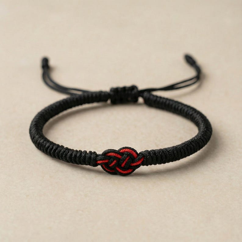 Op de afbeelding: Een zwart gevlochten armband met een rood knoopontwerp in het midden. De armband heeft verstelbare zwarte koorden voor de maatvoering. De achtergrond is een neutraal beige.