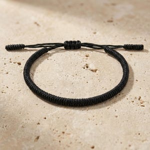 Può includere: Un braccialetto nero intrecciato con chiusura a nodo scorrevole regolabile. Il braccialetto è realizzato in un materiale spesso e tessuto e presenta un design semplice e minimalista. Il braccialetto è mostrato su una superficie neutra e strutturata.