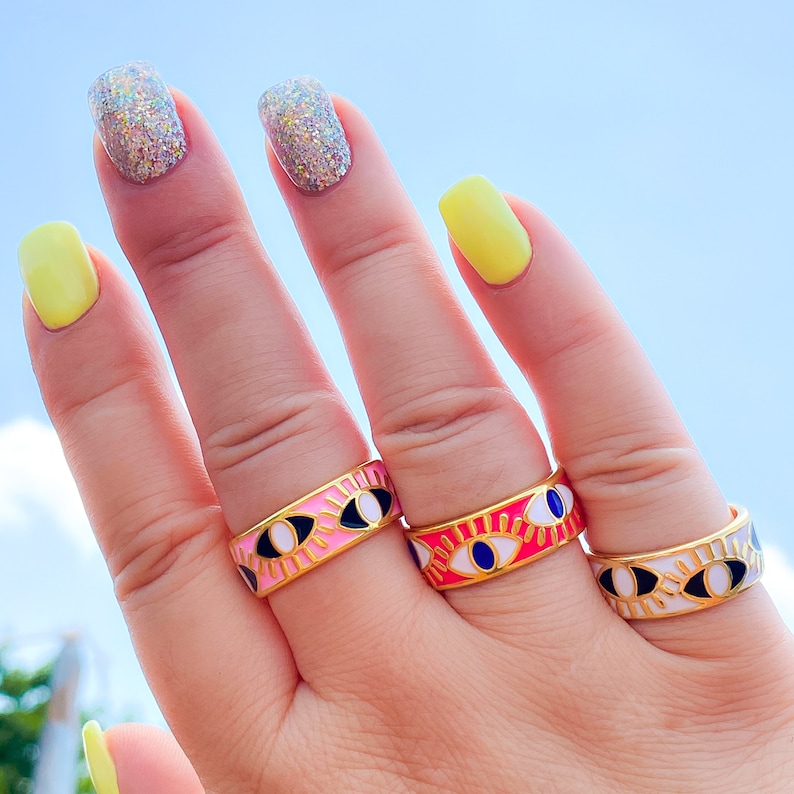 Puede incluir: Tres anillos dorados con dise&ntilde;o de ojo se muestran en una mano contra un cielo azul. Los anillos presentan esmalte rosa, rojo y blanco con detalles de ojos negros y azules. Las u&ntilde;as est&aacute;n pintadas con esmalte amarillo y brillante.