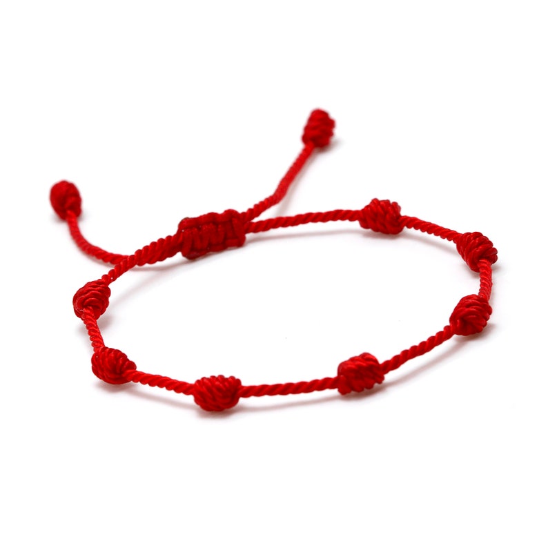 Op de afbeelding: Een rode touw armband met zeven knopen en een verstelbare schuifknoopsluiting. De armband is gemaakt van een dun, geweven rood koord, gepresenteerd tegen een witte achtergrond.