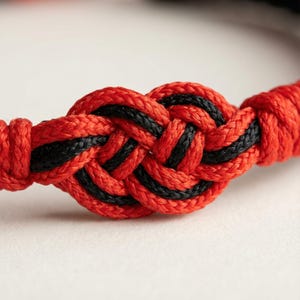 Op de afbeelding: Close-up van een rood en zwart geweven armband. De armband heeft een centraal knoopontwerp met afwisselend rood en zwart touw. De achtergrond is een zachte, neutrale kleur, die de ingewikkelde details van het geweven patroon benadrukt.