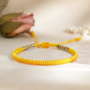 Può includere: Un braccialetto in corda intrecciata giallo con un nodo all'estremità. Il braccialetto è realizzato con una corda spessa e resistente e ha un design semplice ed elegante.