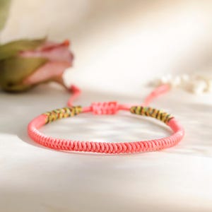 Könnte beinhalten: Ein korallenfarbenes gewebtes Armband mit dekorativen Akzenten. Das Armband verfügt über einen Schiebeknotenverschluss und ist mit gelb-grünen gemusterten Perlen verziert. Das Armband wird auf einer weißen Oberfläche mit unscharfem Hintergrund präsentiert.