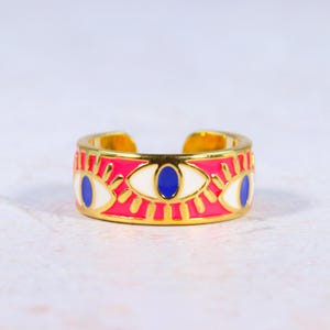 Puede incluir: Un anillo ajustable dorado con una banda rosa con dise&ntilde;os de ojos blancos y azules. El anillo tiene un aspecto brillante y colorido, con un acabado de esmalte detallado. El dise&ntilde;o abierto permite el ajuste de tama&ntilde;o.