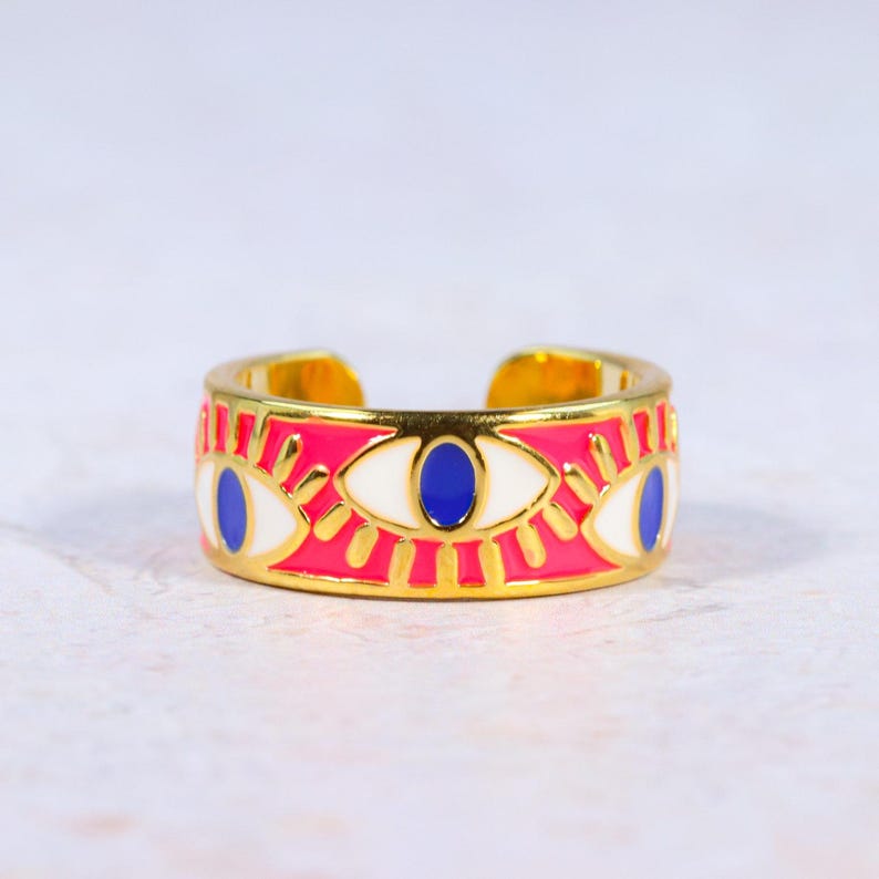Puede incluir: Un anillo ajustable dorado con una banda rosa con dise&ntilde;os de ojos blancos y azules. El anillo tiene un aspecto brillante y colorido, con un acabado de esmalte detallado. El dise&ntilde;o abierto permite el ajuste de tama&ntilde;o.