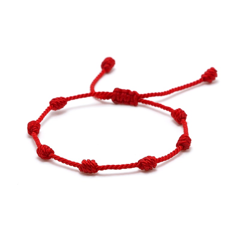 Op de afbeelding: Een rode koordarmband met meerdere knopen. De armband is verstelbaar en heeft een schuifknoopsluiting. De knopen zijn gelijkmatig verdeeld over het rode koord, wat een eenvoudig maar elegant ontwerp cre&euml;ert. De armband wordt tegen een witte achtergrond getoond.