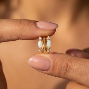 Könnte beinhalten: Ein Paar goldene Creolen mit einem zentralen, marquise-geschliffenen Opal, der von zwei kleinen, runden, klaren Steinen flankiert wird. Die Ohrringe bestehen aus einem polierten Goldmetallband und verfügen über einen sicheren Klappverschluss.