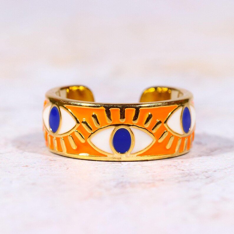 Puede incluir: Anillo ajustable dorado con una banda naranja con un dise&ntilde;o de ojo repetido. Los ojos tienen iris blancos y pupilas azules, con detalles dorados. El anillo est&aacute; abierto en la parte superior.