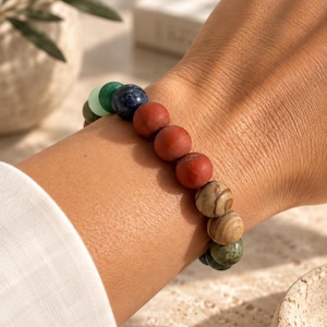 Könnte beinhalten: Ein Perlenarmband bestehend aus einer Reihe runder, polierter Steine in verschiedenen Farben, darunter tiefes Rot, Dunkelblau, Waldgrün, helles Minzgrün und erdiges Braun mit natürlicher Maserung, aufgereiht auf einem elastischen Band.