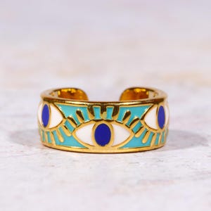 Puede incluir: Anillo ajustable dorado con una banda turquesa con dise&ntilde;os de ojos blancos y azules. El anillo tiene un patr&oacute;n detallado y un acabado pulido, adecuado para el uso diario.