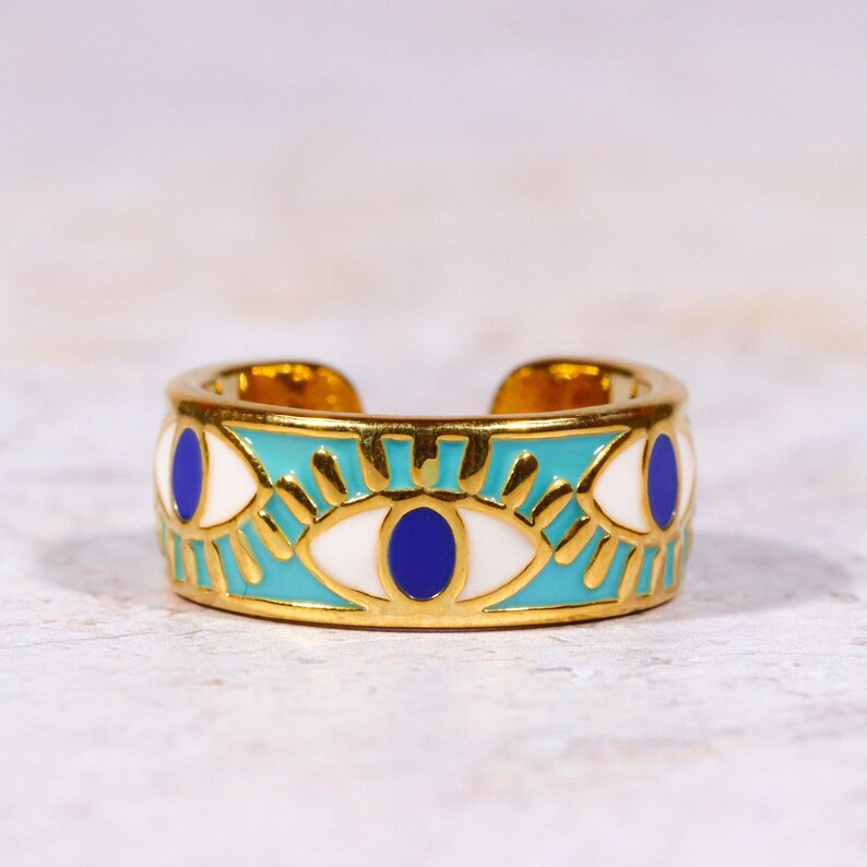 Puede incluir: Anillo ajustable dorado con una banda turquesa con dise&ntilde;os de ojos blancos y azules. El anillo tiene un patr&oacute;n detallado y un acabado pulido, adecuado para el uso diario.
