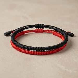 Può includere: Due braccialetti abbinati, uno nero e uno rosso. Entrambi sono realizzati in cordoncino a trama fitta, con nodi scorrevoli regolabili. I braccialetti sono esposti su uno sfondo neutro.