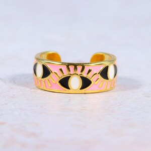 Puede incluir: Anillo ajustable dorado con una banda rosa con un dise&ntilde;o de ojo repetido. Los ojos tienen detalles en blanco y negro, y el anillo tiene un acabado brillante. Es un accesorio de moda.