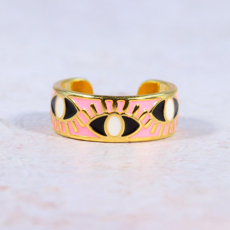 Puede incluir: Anillo ajustable dorado con una banda rosa con un dise&ntilde;o de ojo repetido. Los ojos tienen detalles en blanco y negro, y el anillo tiene un acabado brillante. Es un accesorio de moda.