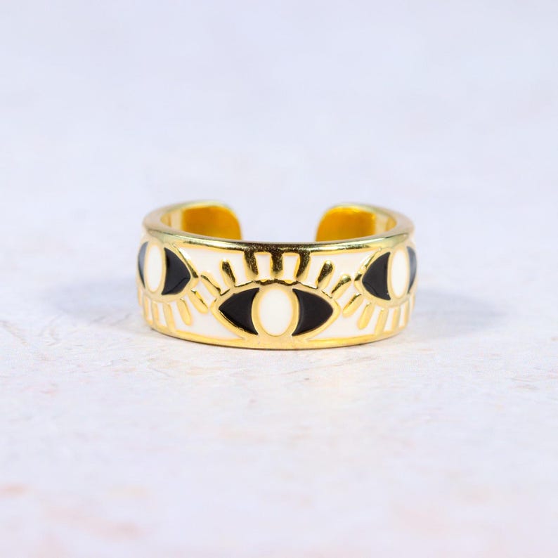 Puede incluir: Anillo ajustable dorado con una banda blanca con dise&ntilde;os de ojos negros y blancos. El anillo tiene un acabado pulido y una est&eacute;tica minimalista. El anillo mide aproximadamente 1,9 cm de ancho, adecuado para el uso diario.