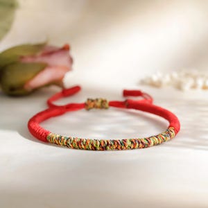 Op de afbeelding: Een rode armband met een geweven, veelkleurig middengedeelte. De armband is verstelbaar met rode koorden en een kleine, decoratieve sluiting. De achtergrond is een zachte, neutrale kleur.